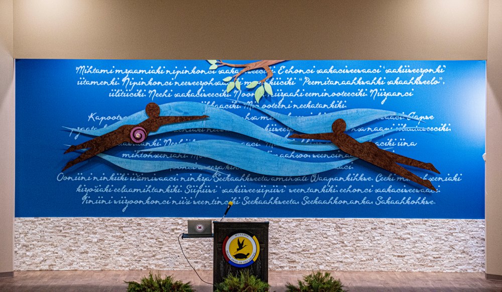 Two dark figures in water swimming toward a tree branch over the story Where the Miamis First Came From written in the Myaamia language.