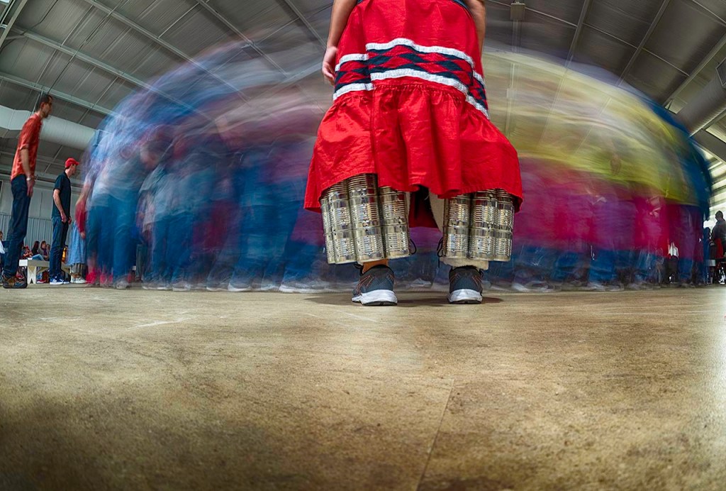 A woman wearing shaker cans watches the blurred stomp dance circle in a timelapse photo