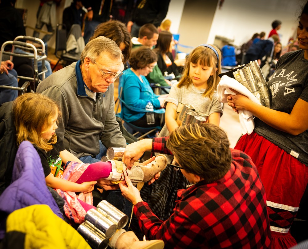 A young girl receives help from her father in lacing her shaker cans while her family watches