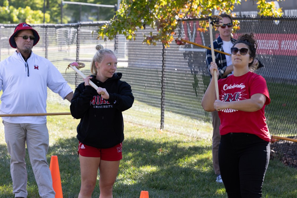 Two women standing with wooden Myaamia lacrosse sticks over their shoulders as they prepare to launch a ball towards the target. 