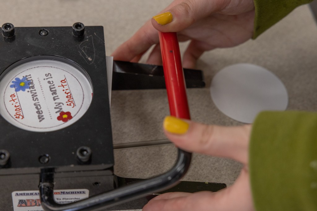 A person wearing yellow nail polish uses both thumbs to press a lever to seal a plastic coating over a button that reads "Sharita weenswiaani, My name is Sharita." 