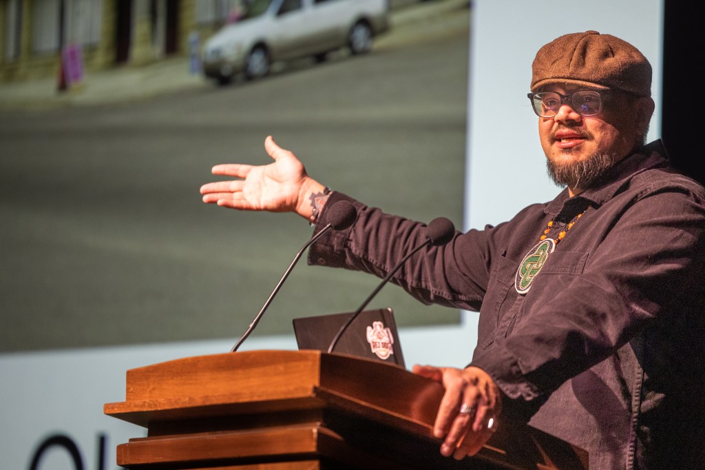 Sterlin Harjo stands at a podium wearing a brown hat and shirt with his arm outstretched towards a screen out of the shot. 