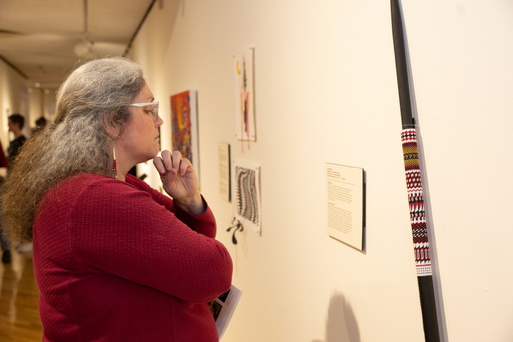 A woman in a red sweater reads an artist statement next to a black spear with rows of red, black and yellow beads covering it.