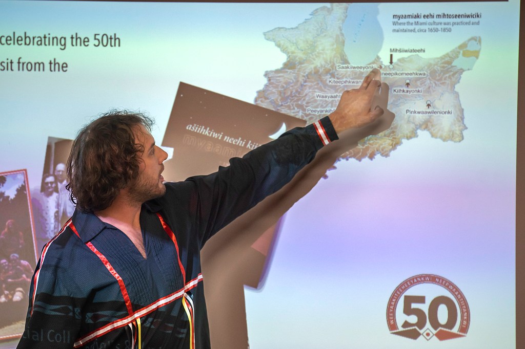 A man wearing a black shirt with red ribbons, points to a map of the Myaamia homelands projected on a screen.