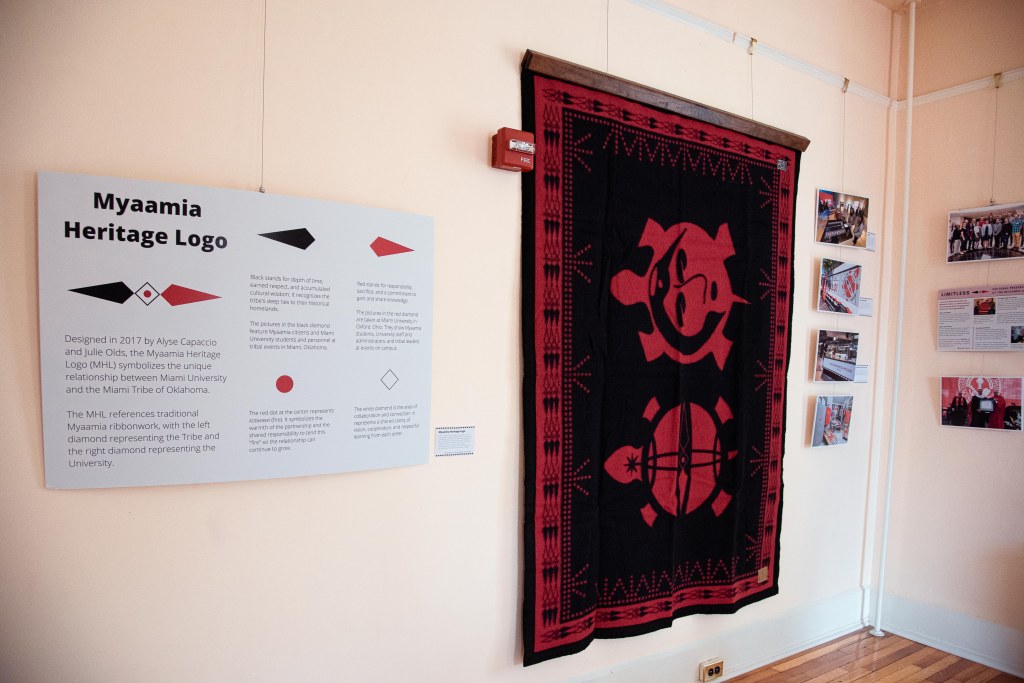 A black and red wool blanket showing two turtle designs hangs on the wall next to a graphic detailing the different aspects of the Myaamia Heritage Logo.