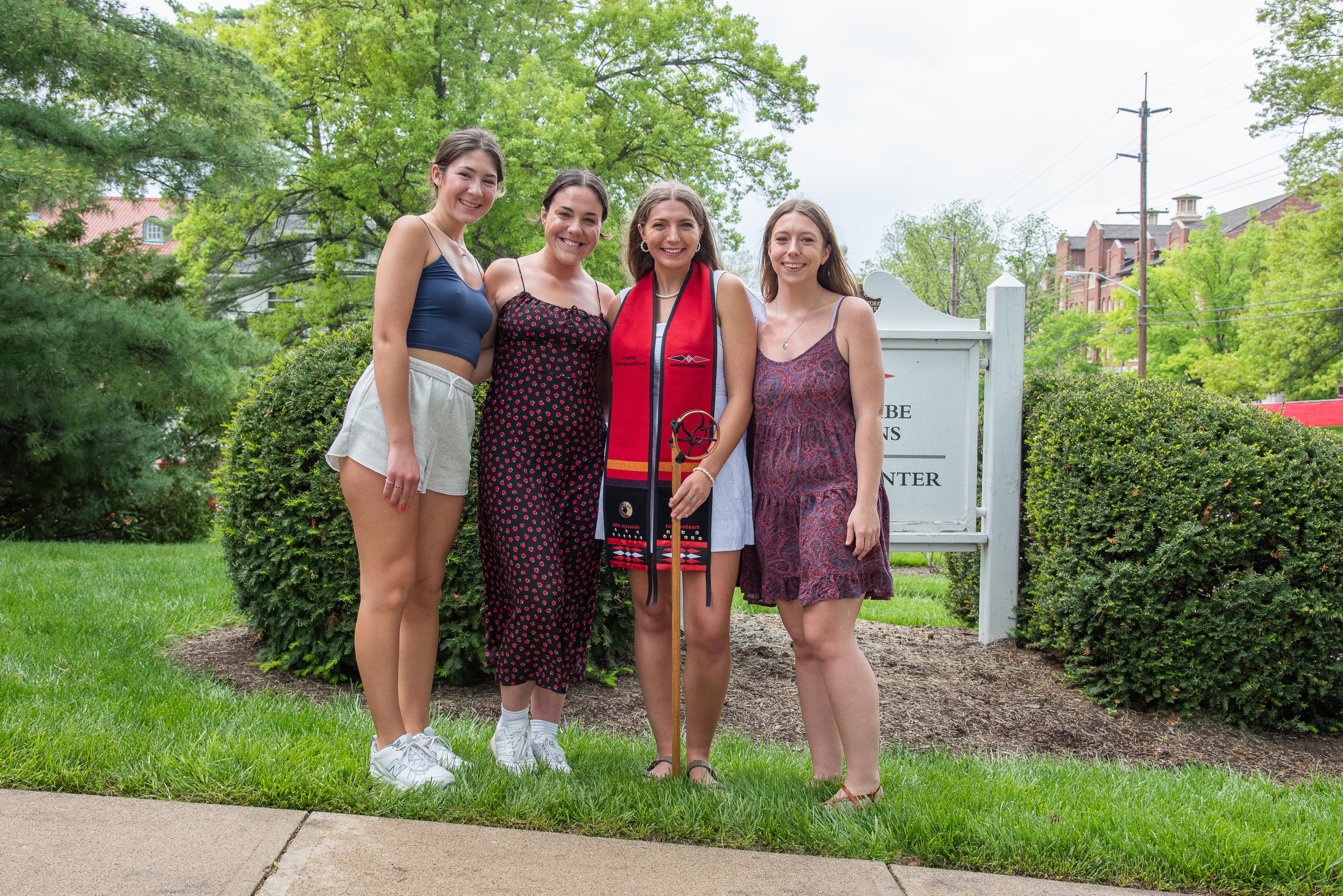 Four young women stand outside, the graduate stands in the middle holding her wooden lacrosse stick.