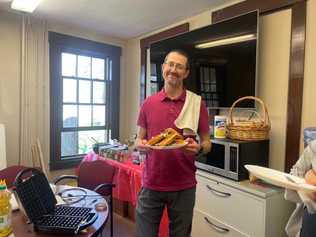 A man smiles for the camera, holding a plate of fresh waffles with a kitchen towel thrown over his shoulder. 