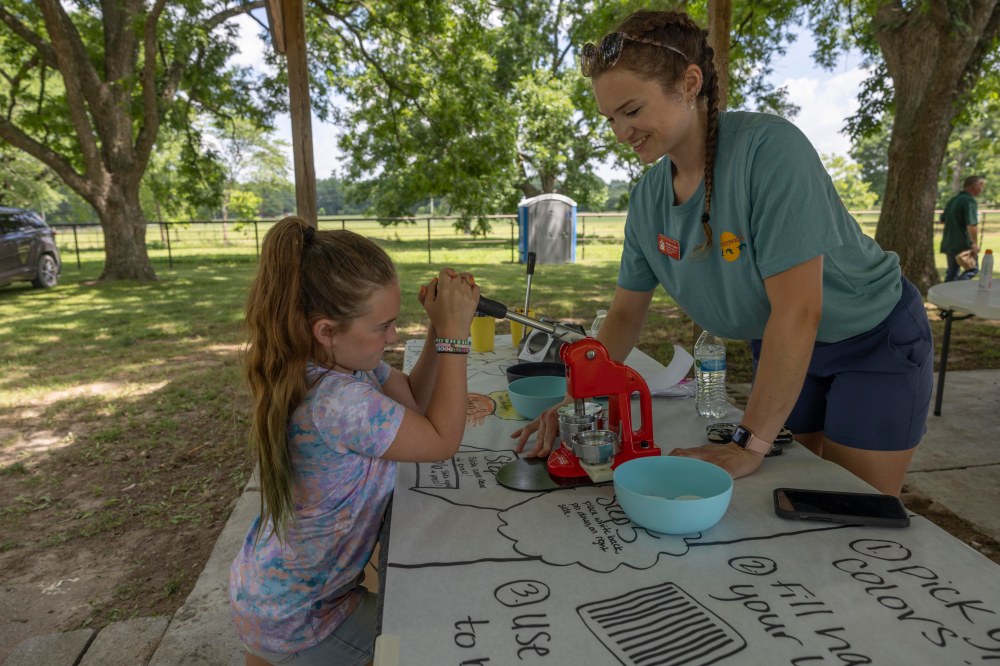 A child pulling the lever of a button maker while an adult supports the machine