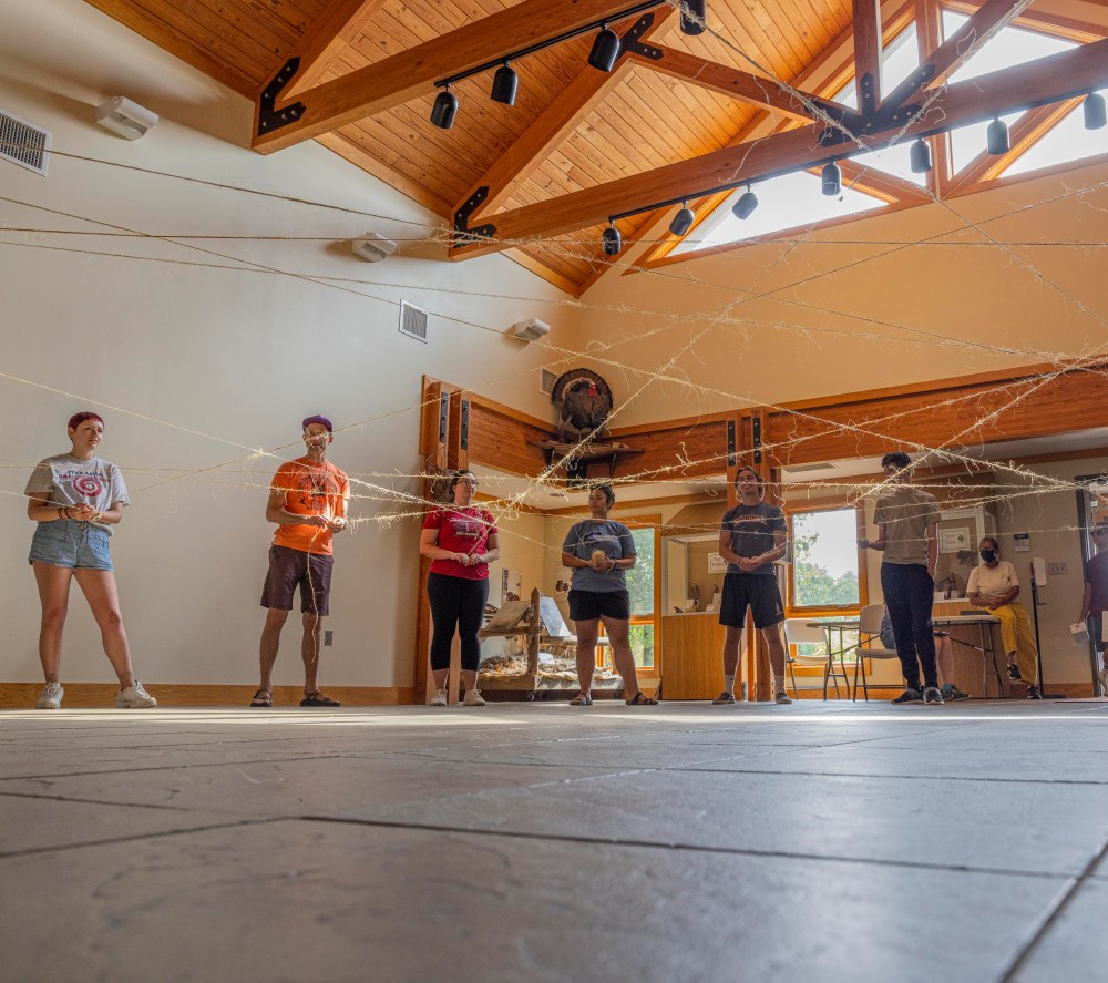 A group of people stand in a circle each holding a piece of twine, forming a web in the middle of the circle.