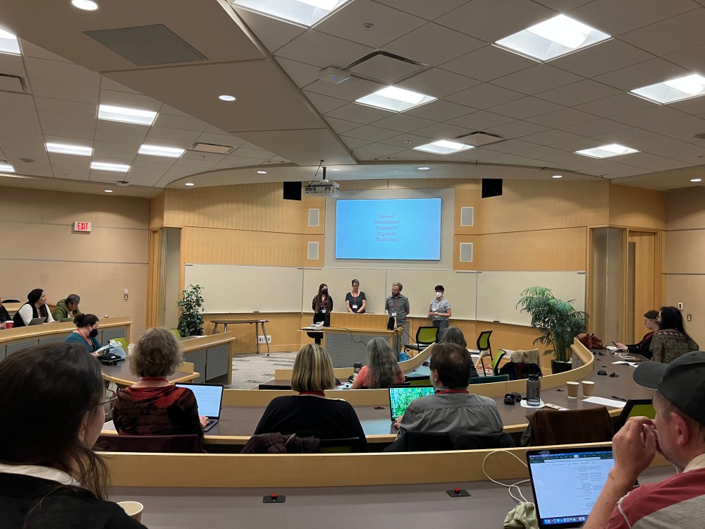 Four people standing at the front of an audience in auditorium style seating