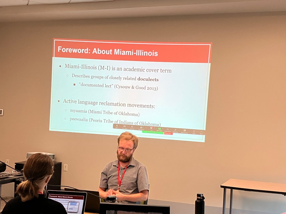 Man sitting in front of a projector screen addressing the audience