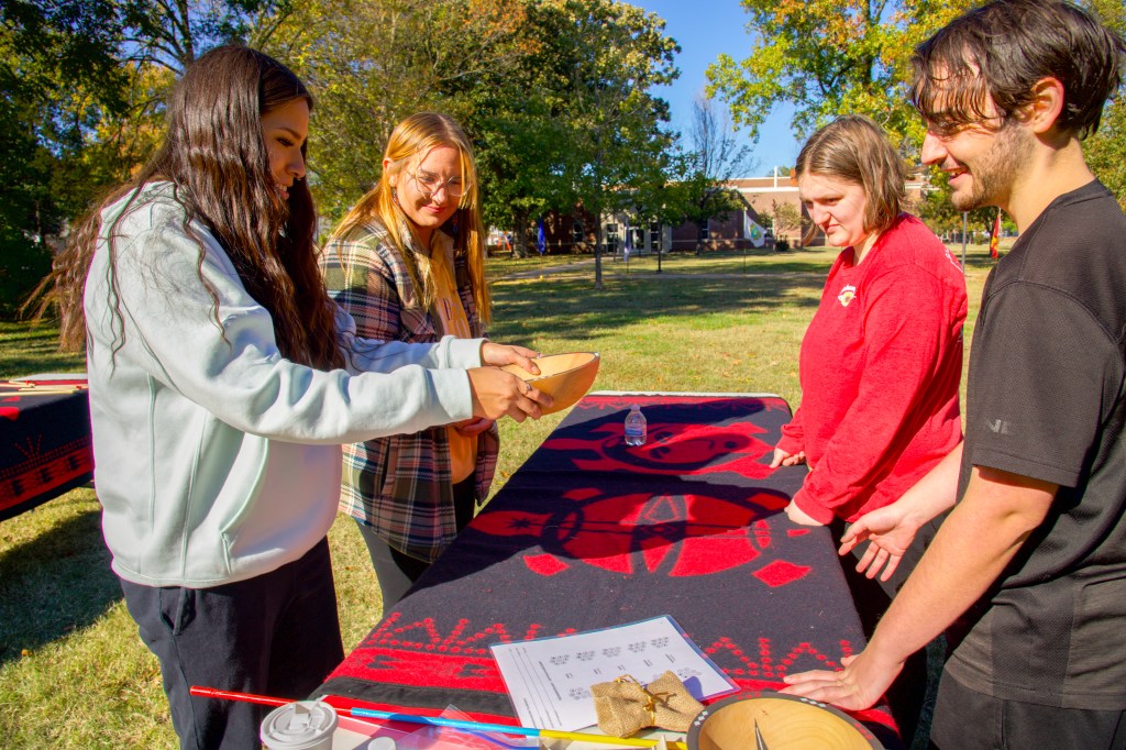 A man and three women stand around a table watching as one woman tosses bowl game pieces in a wooden bowl