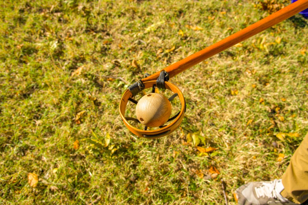 A wooden lacrosse ball with several holes drilled in it sitting in the round net of a wooden lacrosse stick