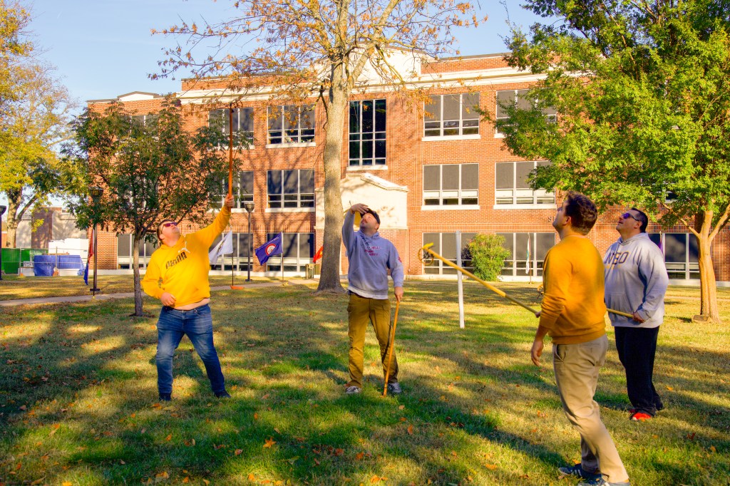 Four men look up at a tossed lacrosse ball with their wooden lacrosse sticks ready