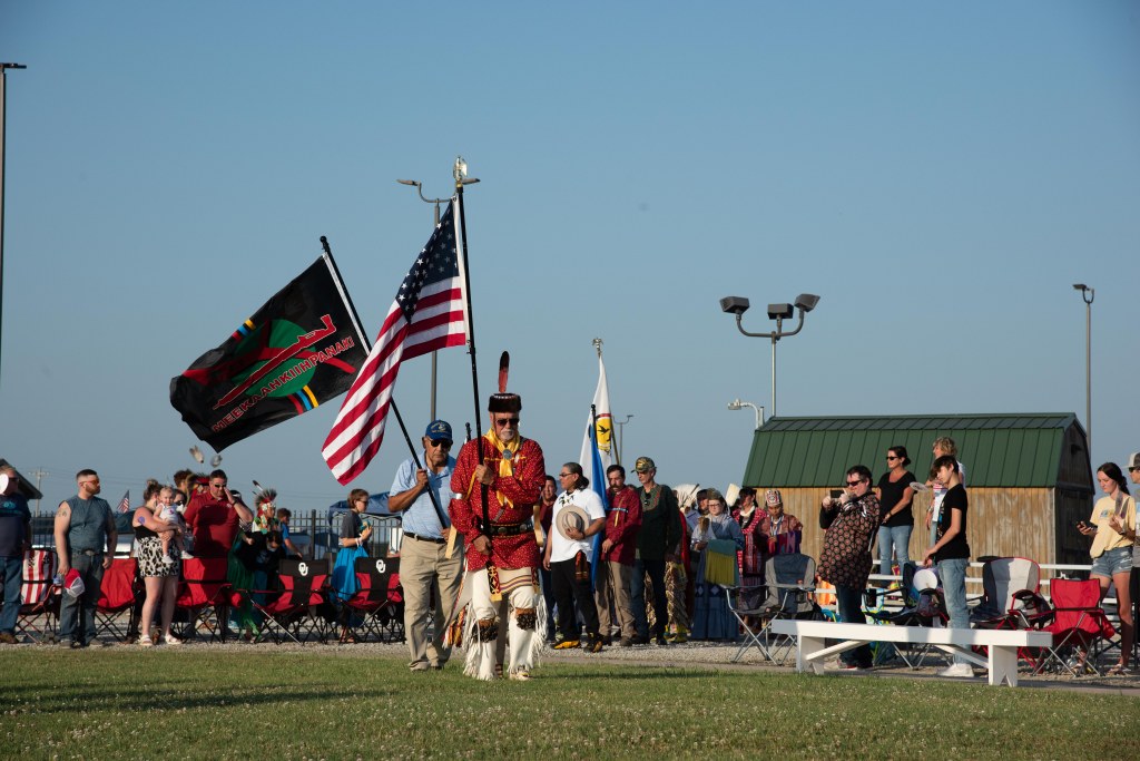A photo of two dancers carrying flags entering the Miami Tribe's pow wow ground in Miami, OK. The first dancer carries the American flag and the second carries the Tribe's Veterans Flag. A crowd stands in the background watching.