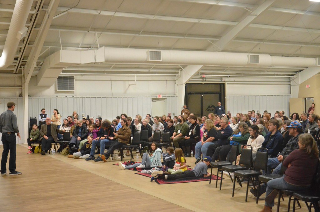 A group of people sit in a semi-circle, watching a man standing in the middle tell a story