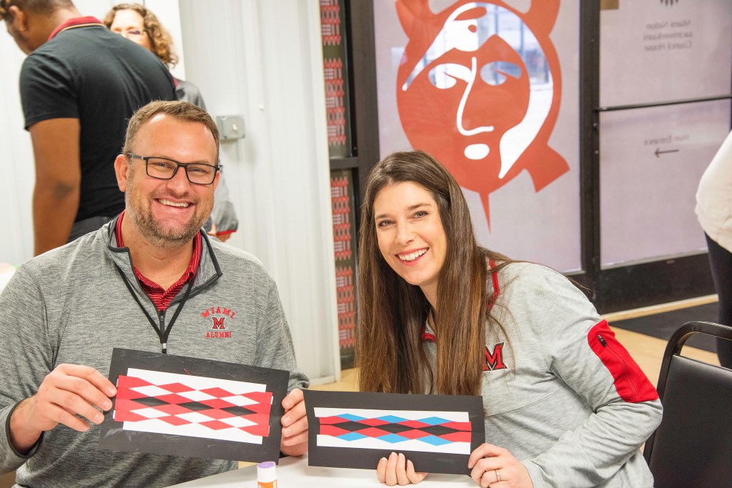A man and a woman sit together holding up their ribbonwork bookmark projects.