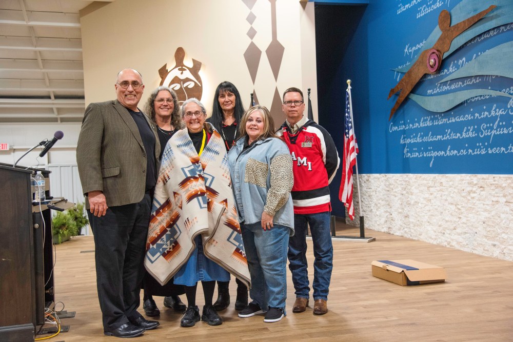 6 people smile for a group photo on stage, the woman in the middle is wrapped in a blanket and wears a medal of honor around her neck