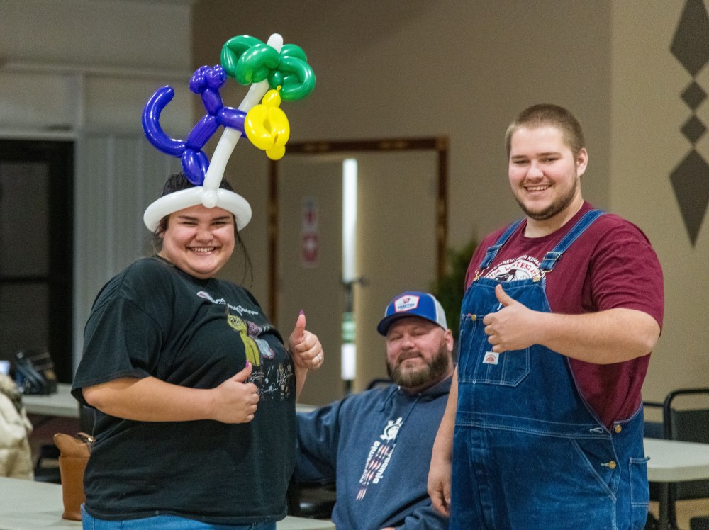 3 people stand together smiling at the camera, one wears a balloon hat