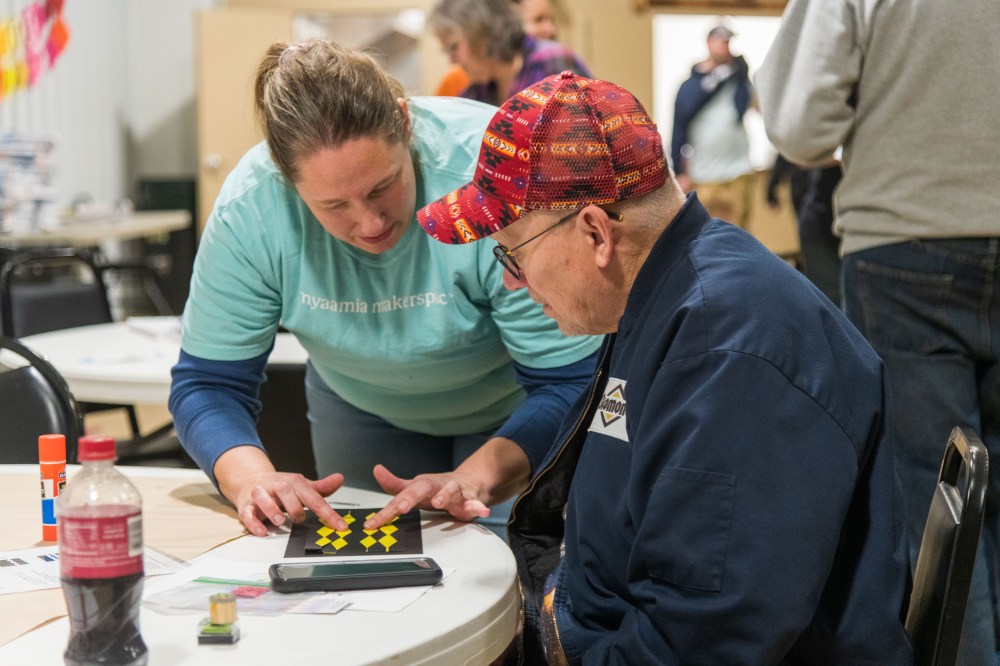 A man sits a table with a bookmark craft in front of him while a woman presses on the bookmark to bind the glue