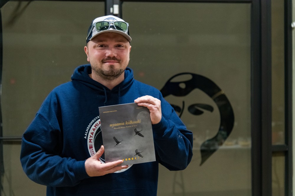 A man smiles at the camera while holding a grey, hardcover book