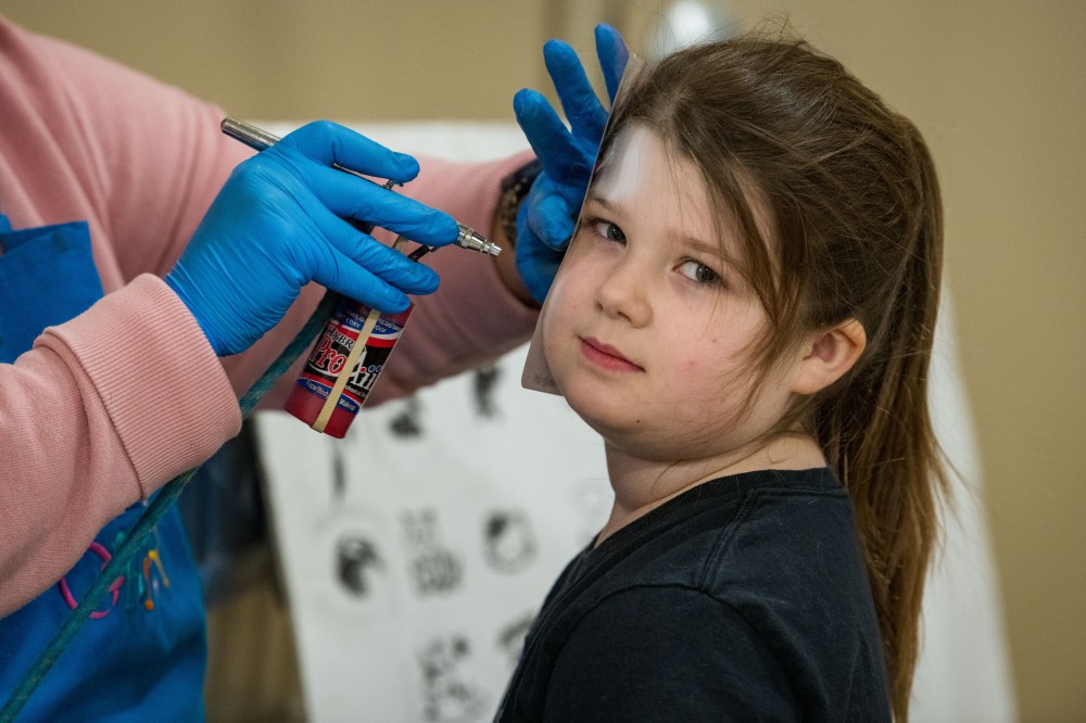 A young child looks at the camera while an adult uses an airbrush to paint a deign on her face