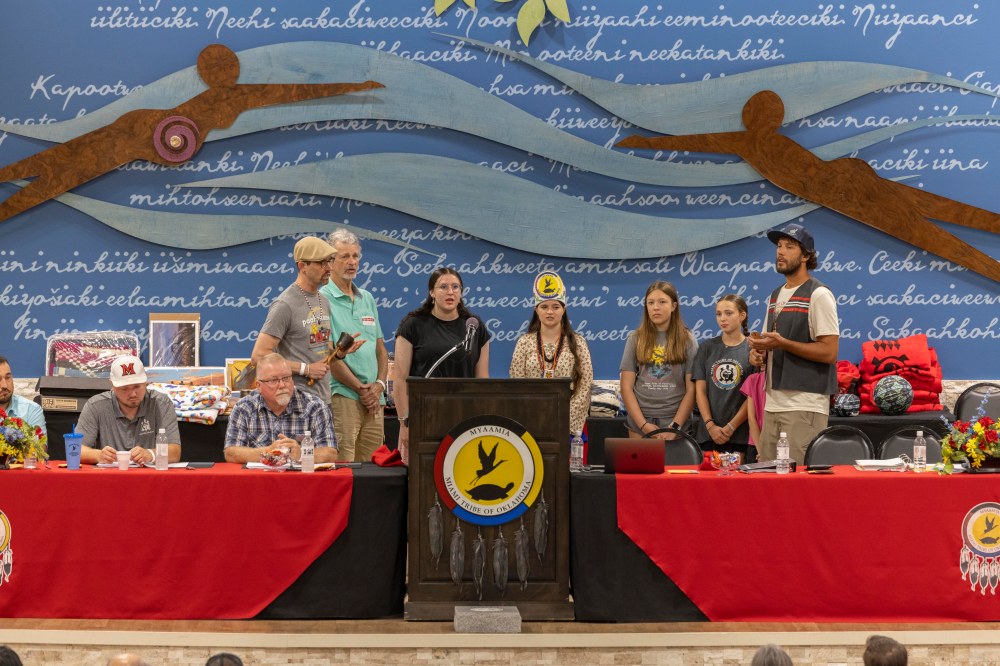 Myaamia adults and youth stand at a podium singing, two of them hold rattles to keep rhythm