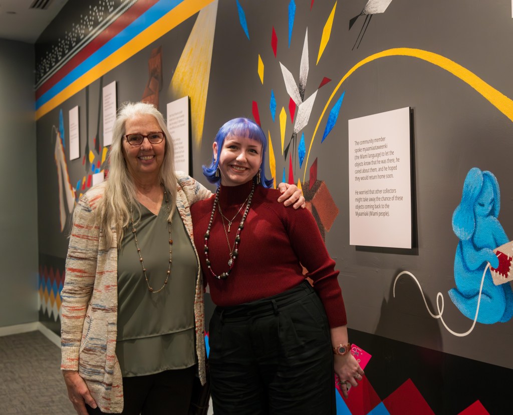 Two women posing in front of a mural at the First Americans Museum
