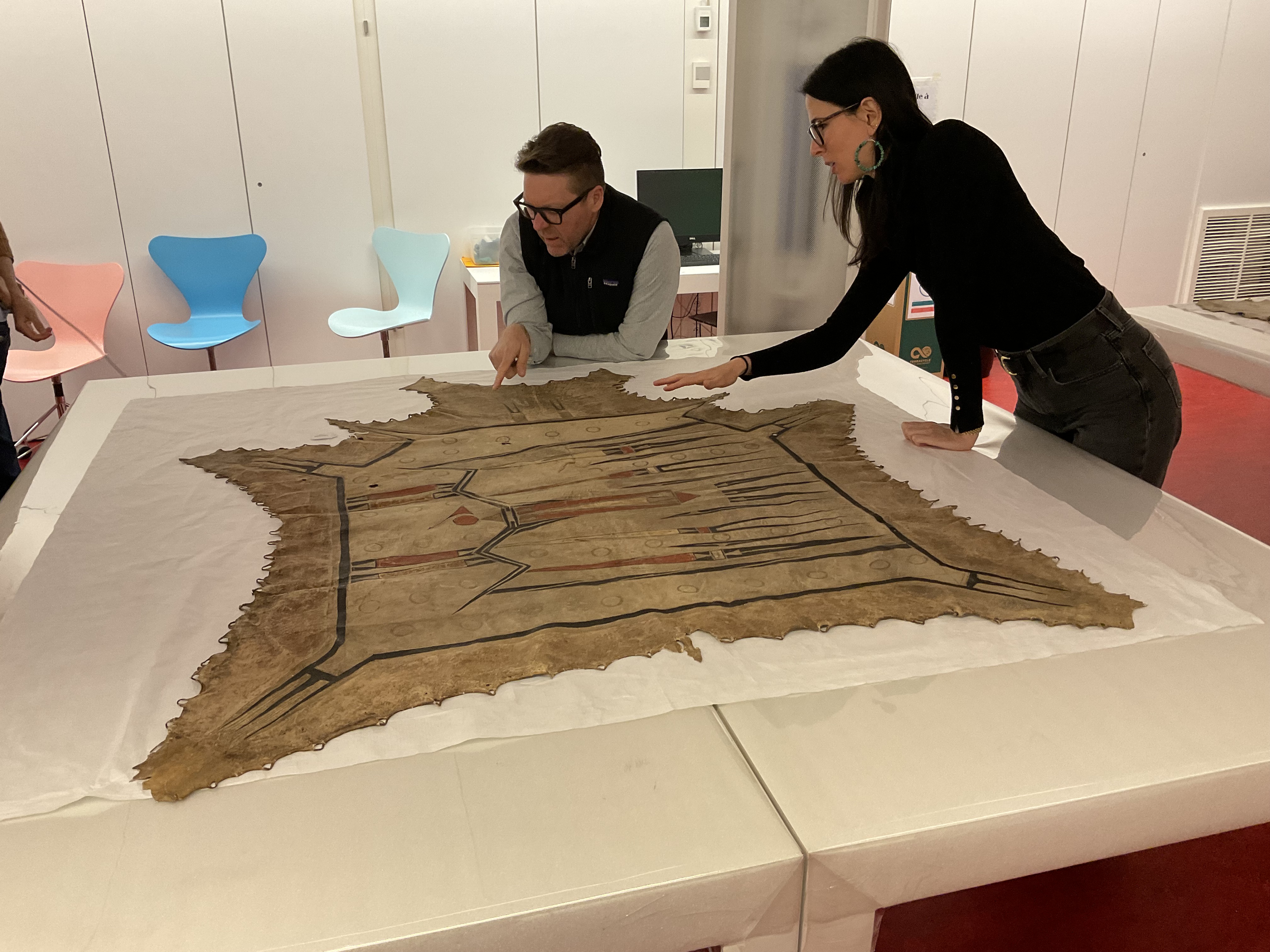 A large painted hide robe spread on a table with a man in the background pointing at a spot close to him and a woman next to him motioning to that same area