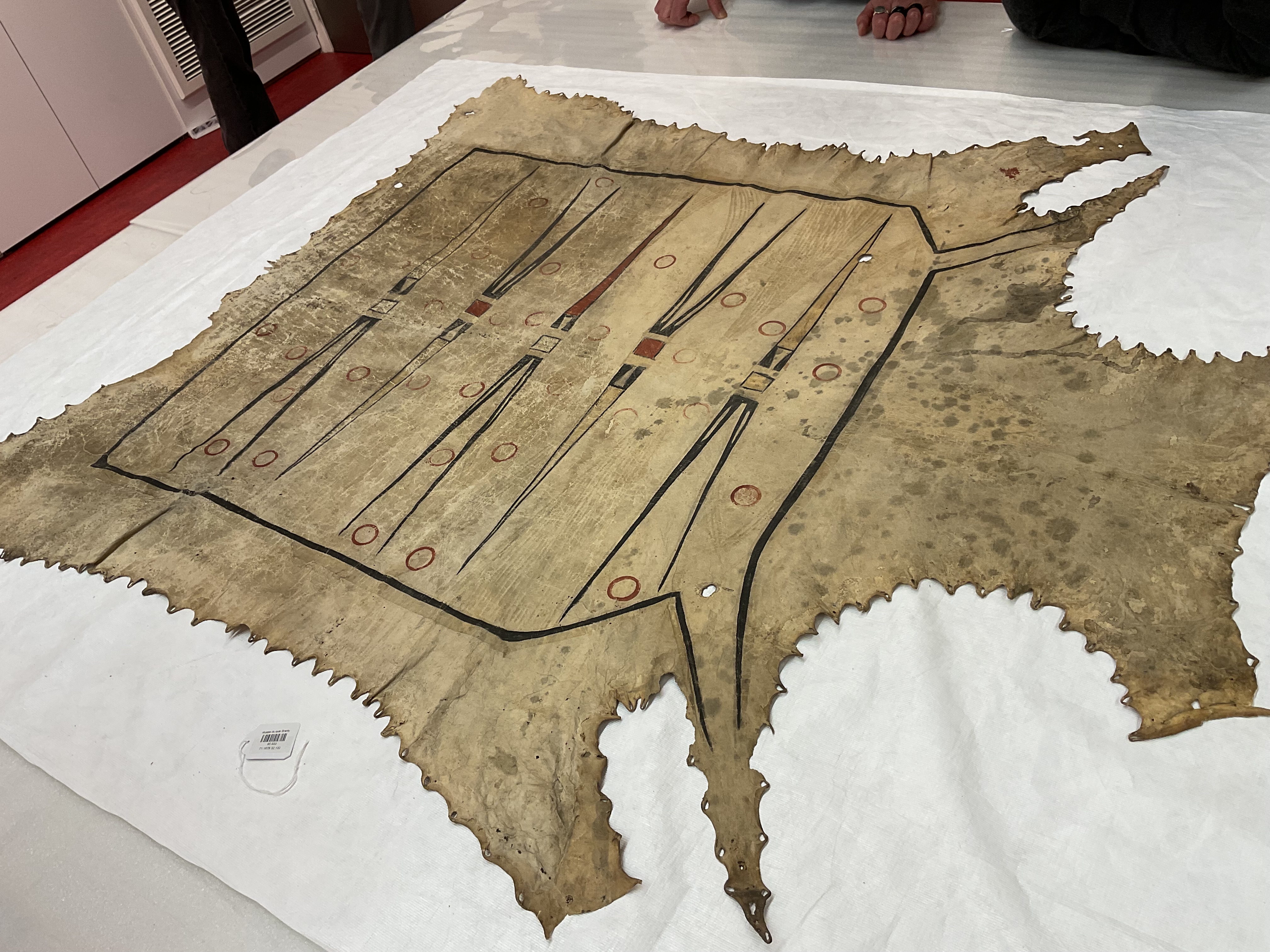 A painted hide robe displayed on a table. The design features elongated triangles and small circles in clear, black, red, and yellow pigments.