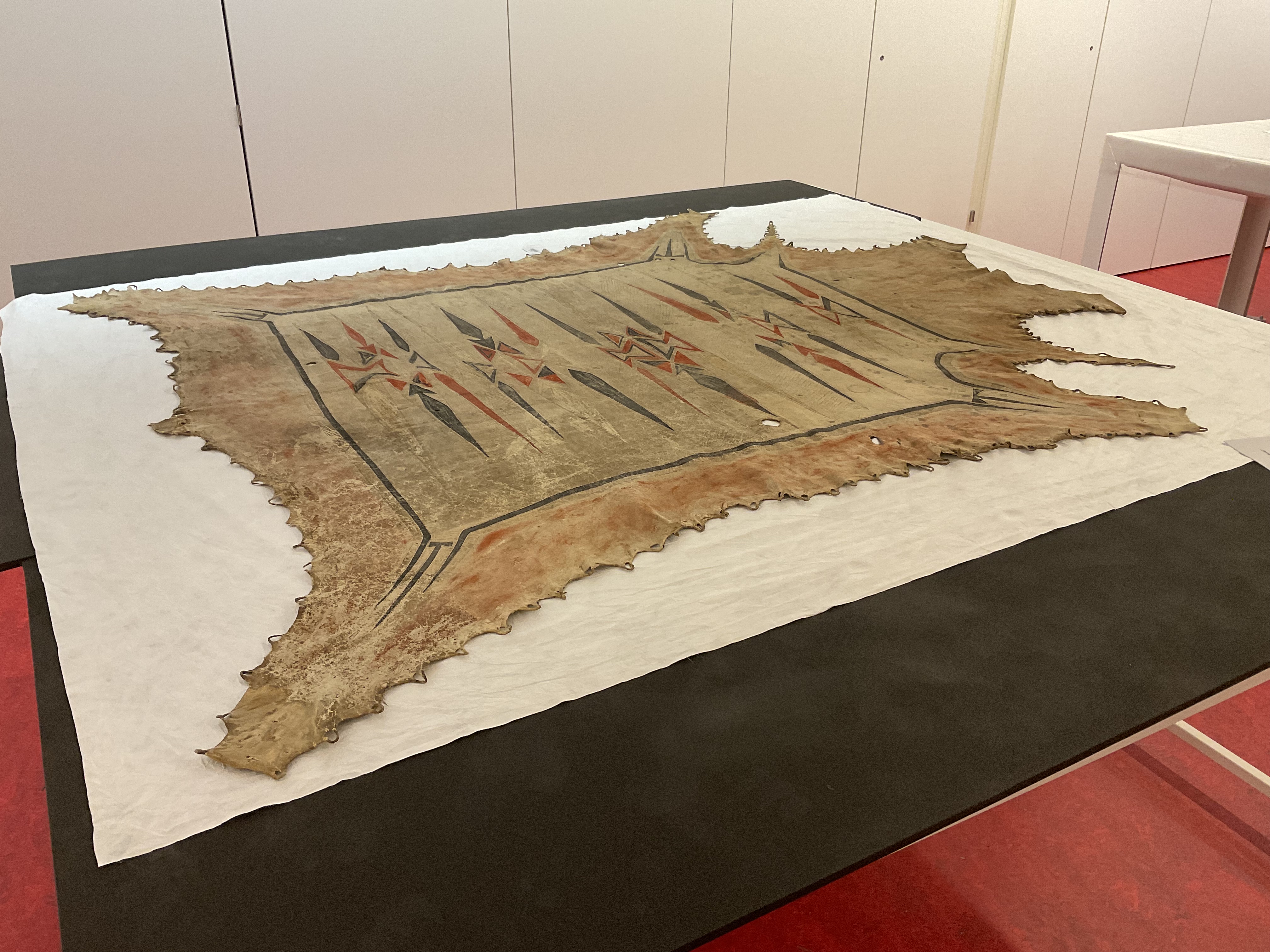 A painted hide robe displayed on a table. The design features elongated diamonds and hourglass shapes in clear, black, and red pigments.
