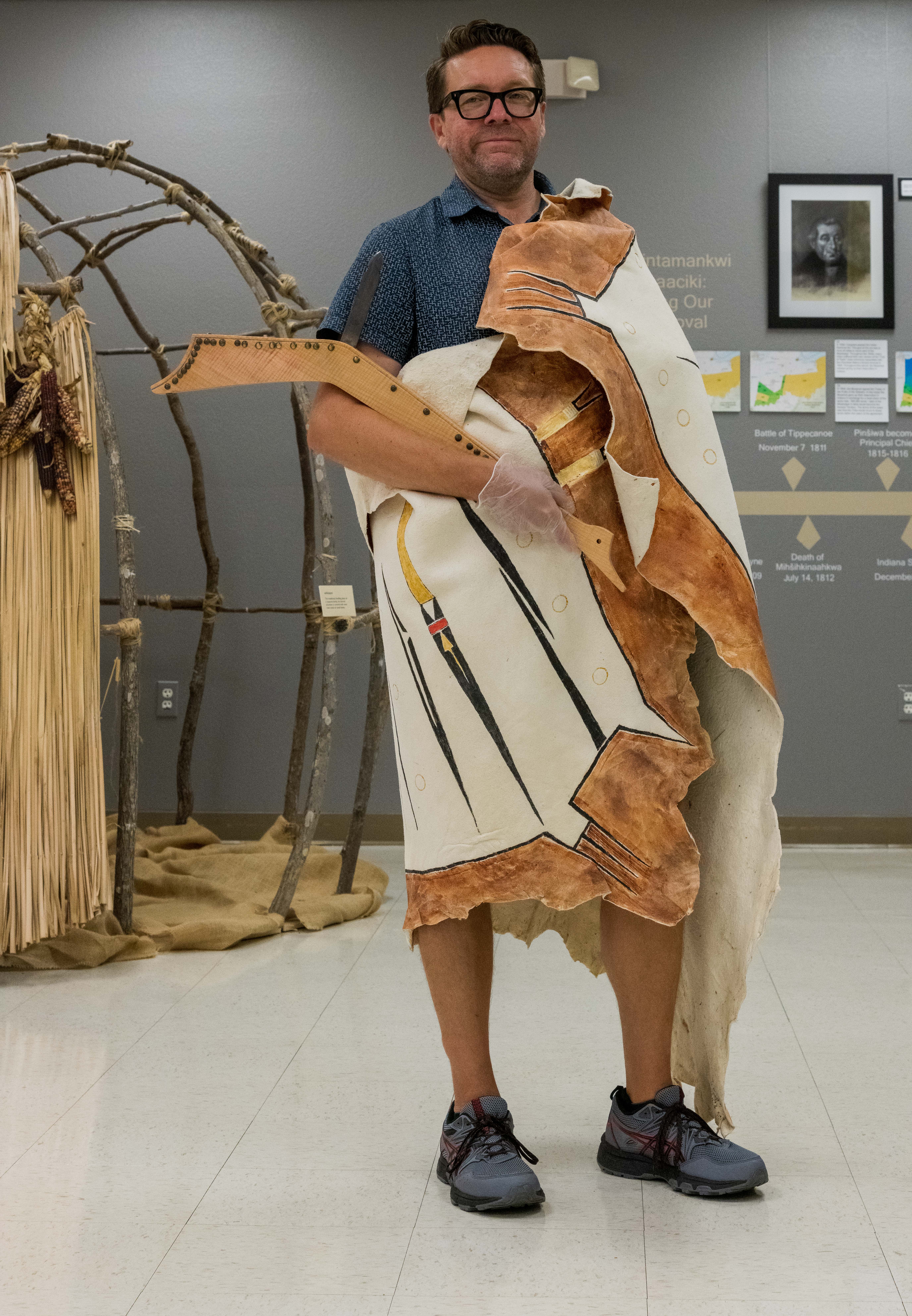 A man wearing a replica painted hide robe. It is draped across his left shoulder, wrapped around his back, and tucked under his right arm. Across his right arm, he is holding a replica gunstock war club.