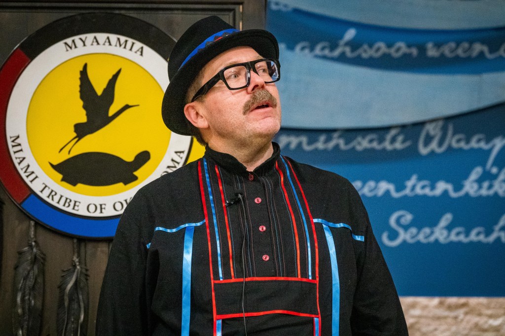 A man wearing a hat with a ribbonwork hat band and ribbon shirt in front of a podium bearing the Miami Tribe of Oklahoma seal
