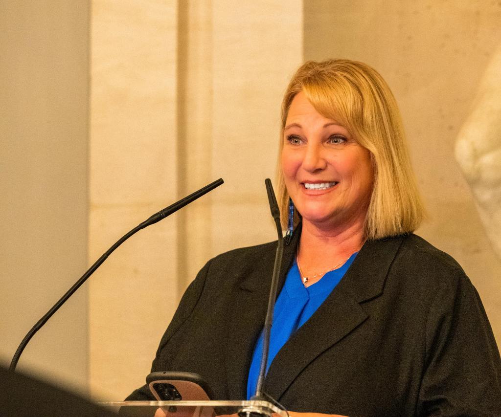 Woman (Chief Rosanna Dobbs) stands at a podium giving a speech. A portion of a statue can be seen behind her.
