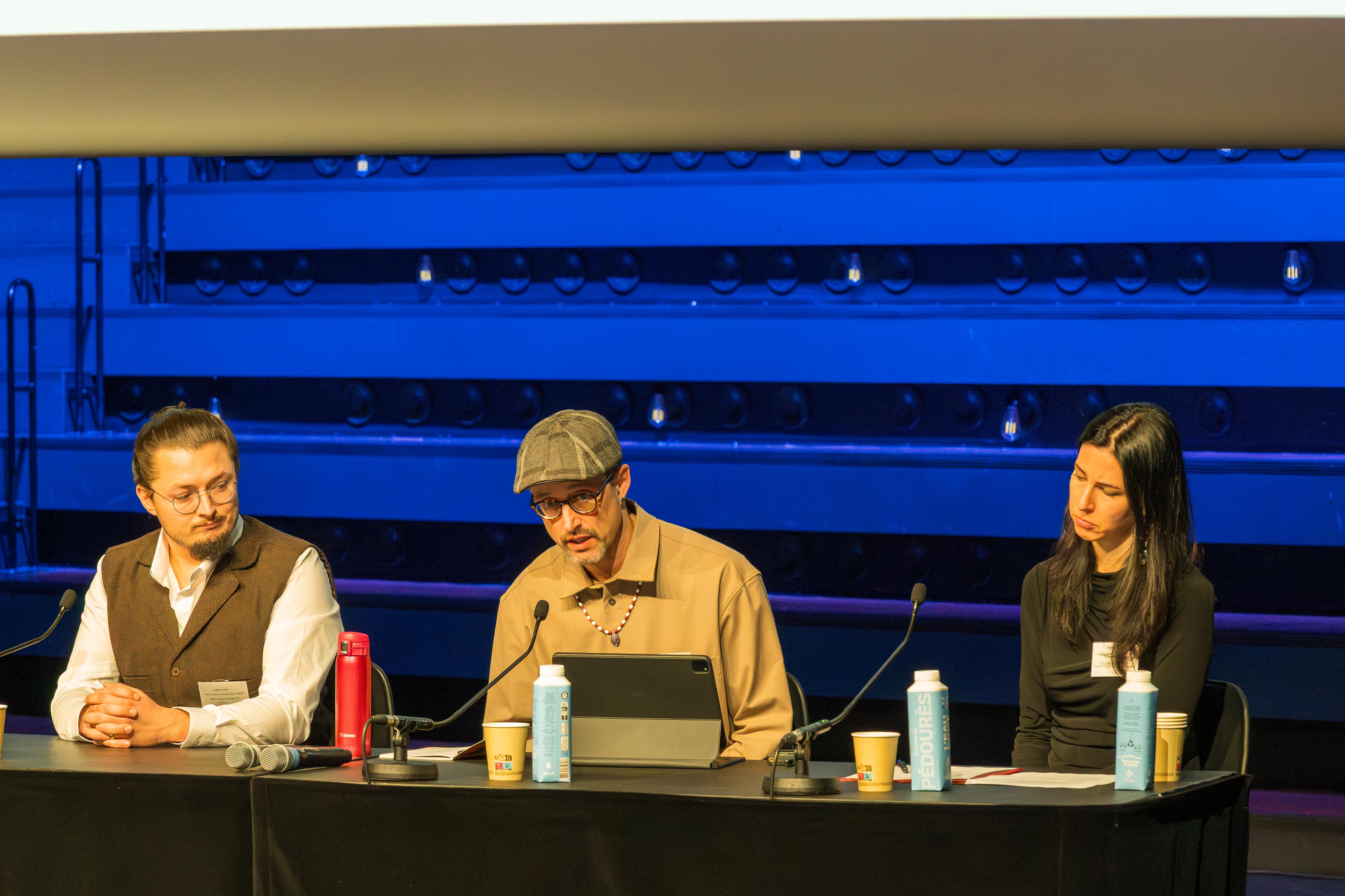 Three presenters sit at a table. The presenter in the middle is speaking.