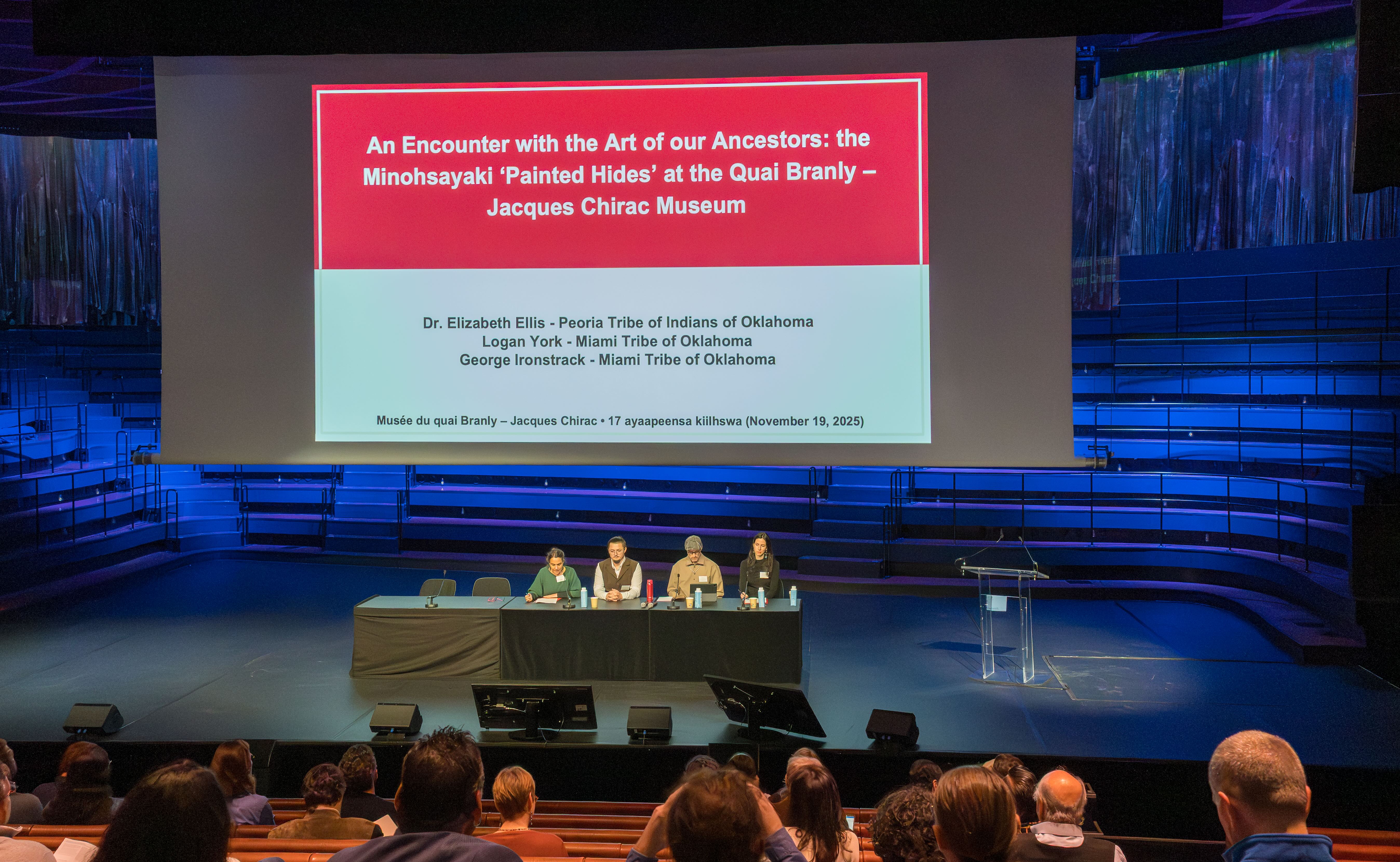 Four speakers sit at a table on a stage beneath a projected slide that reads: "An Encounter with the Art of Our Ancestors: the Minohsayaki 'Painted Hides' at the Quai Branly - Jacques Chirac Museum"